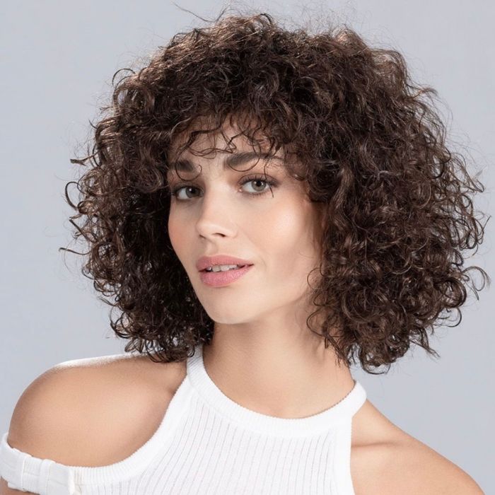 Femme aux yeux clairs portant un débardeur blanc et une perruque brune aux cheveux bouclés, coupe au carré